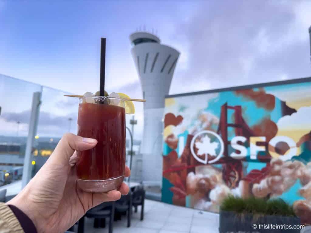 Hand holding up a caesar cocktail on the SFO Air Canada Lounge terrace with a mural of San Francisco in the background. Behind that, the airport control tower