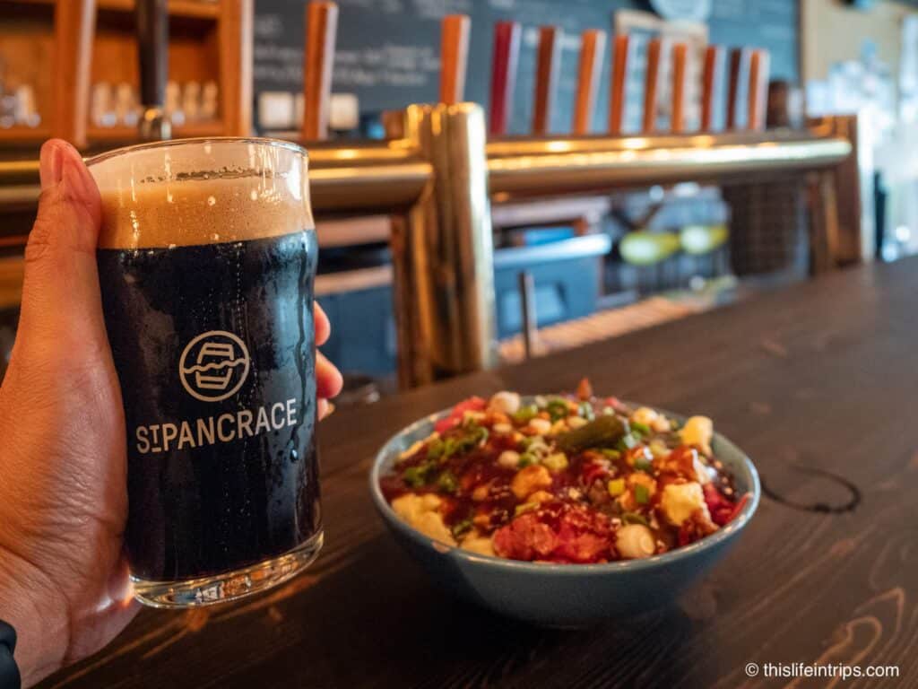 Hand holding pint of dark beer at Saint-Pancrace microbrewery with bowl of poutine in background