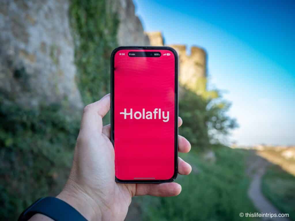 Hand holding up iPhone with Holafly eSIM app splash screen in front of castle in Europe