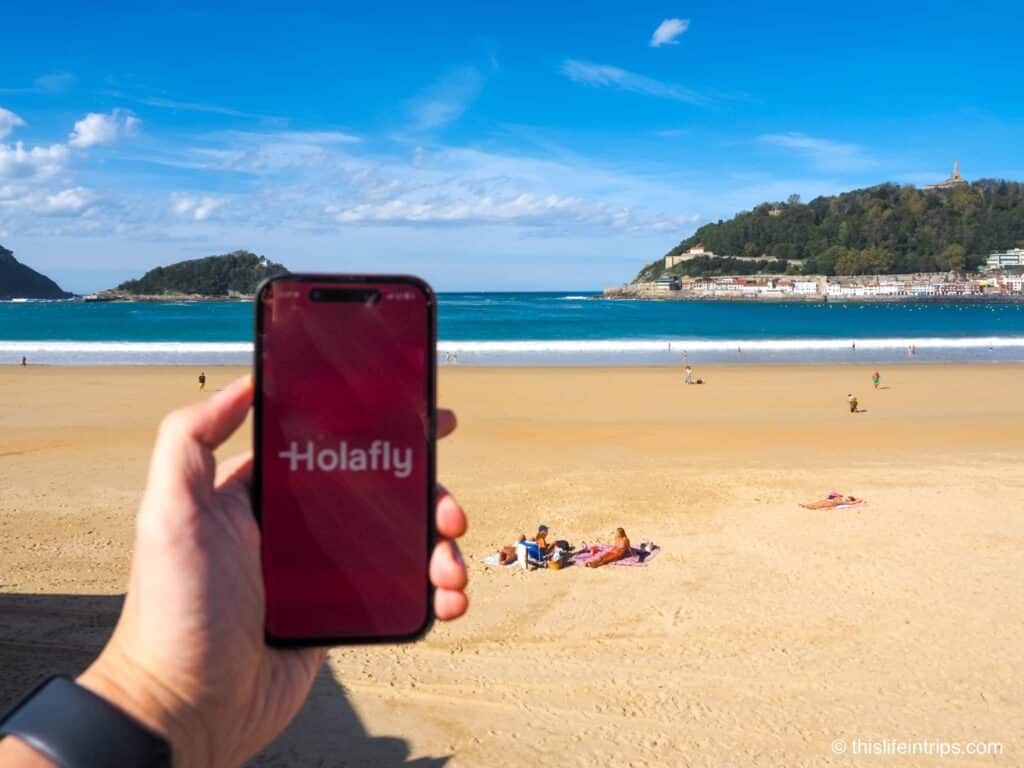 Hand holding up iPhone with Holafly eSIM app splash screen in front of a beach in San Sebastian Spain