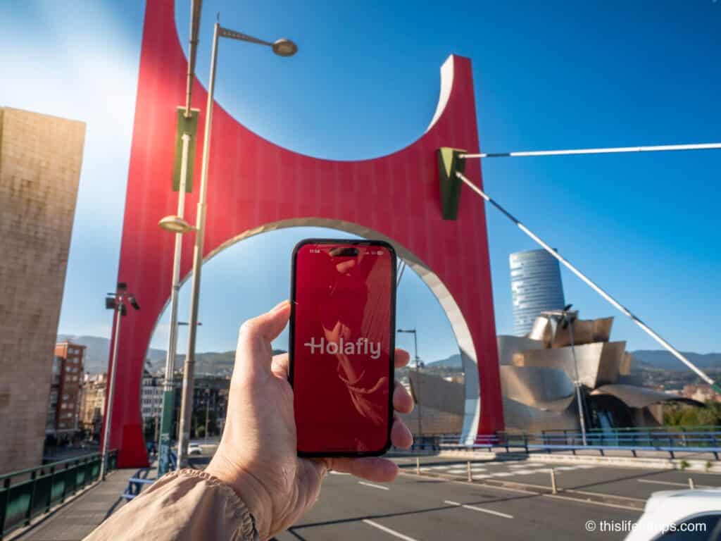 Hand holding up iPhone with Holafly eSIM app splash screen in front of the Guggenheim museum in Bilbao Spain