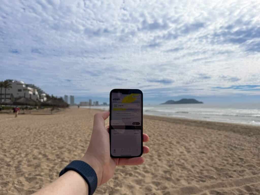 hand holding up iPhone with Saily eSIM app loaded on screen on a beach