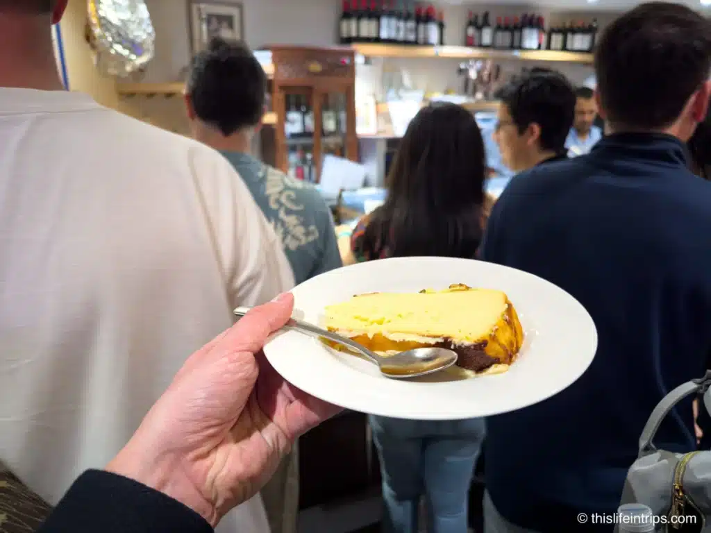 Hand holding up a plate of Basque cheesecake at a crowded tapas bar in San Sebastian, Spain
