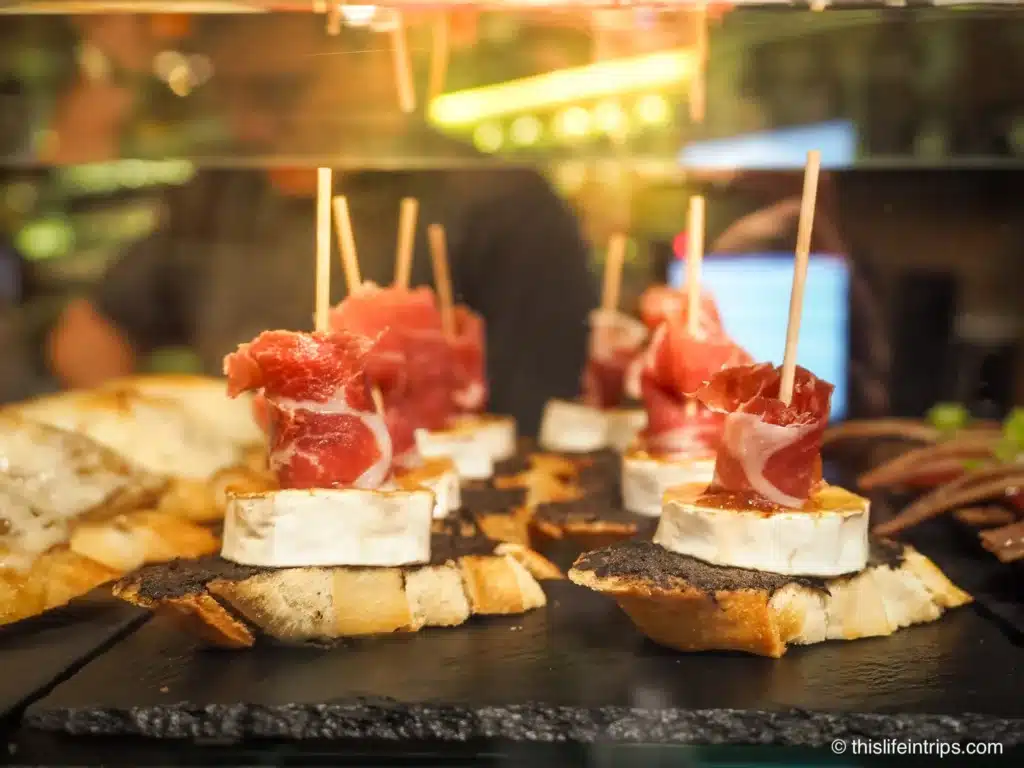 Photo of so,e of the best pintxos in San Sebastian - cured Iberia ham piled on top of a big slab of Brie on slice of baguette held together by a toothpick