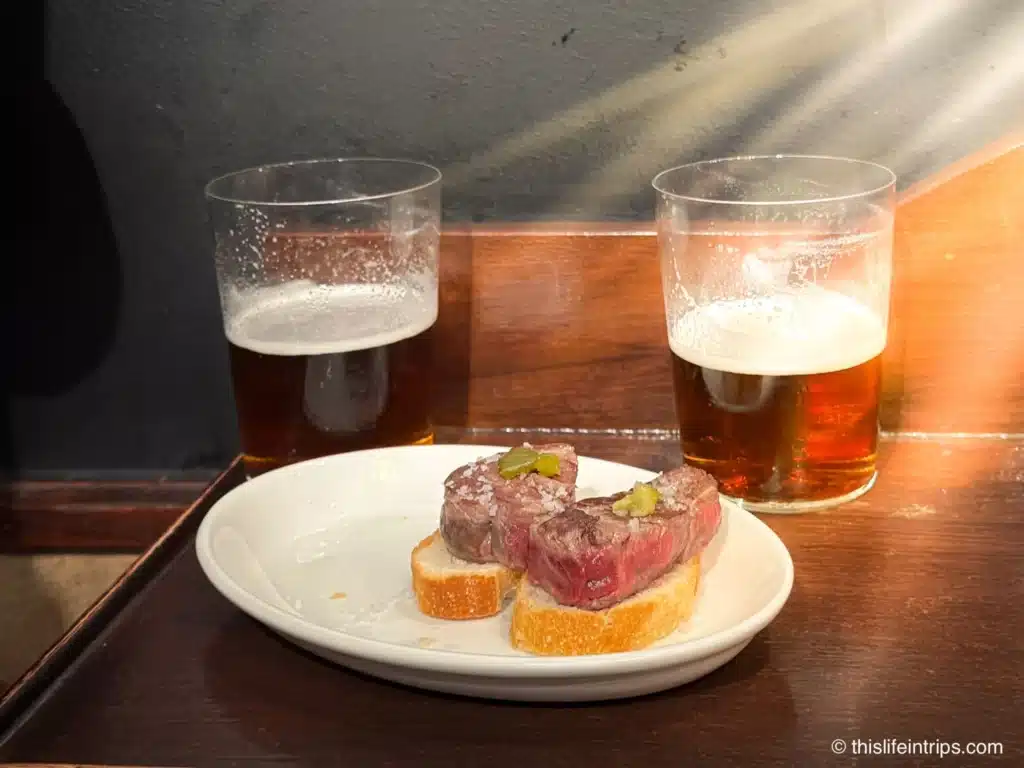 Two pieces of steak on cut up baguettes in front of two half drunk glasses of beer while on San Sebastian food tour