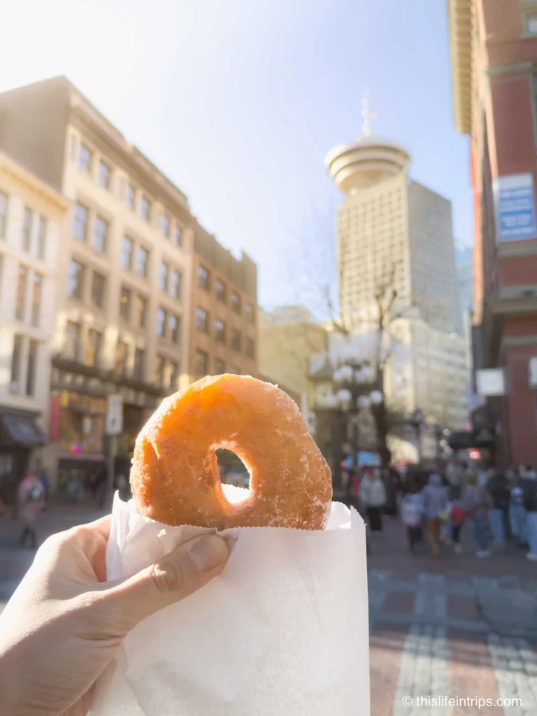 Hand holding up The Honey dip from Lee's Donuts in front of Vancouver steam clock